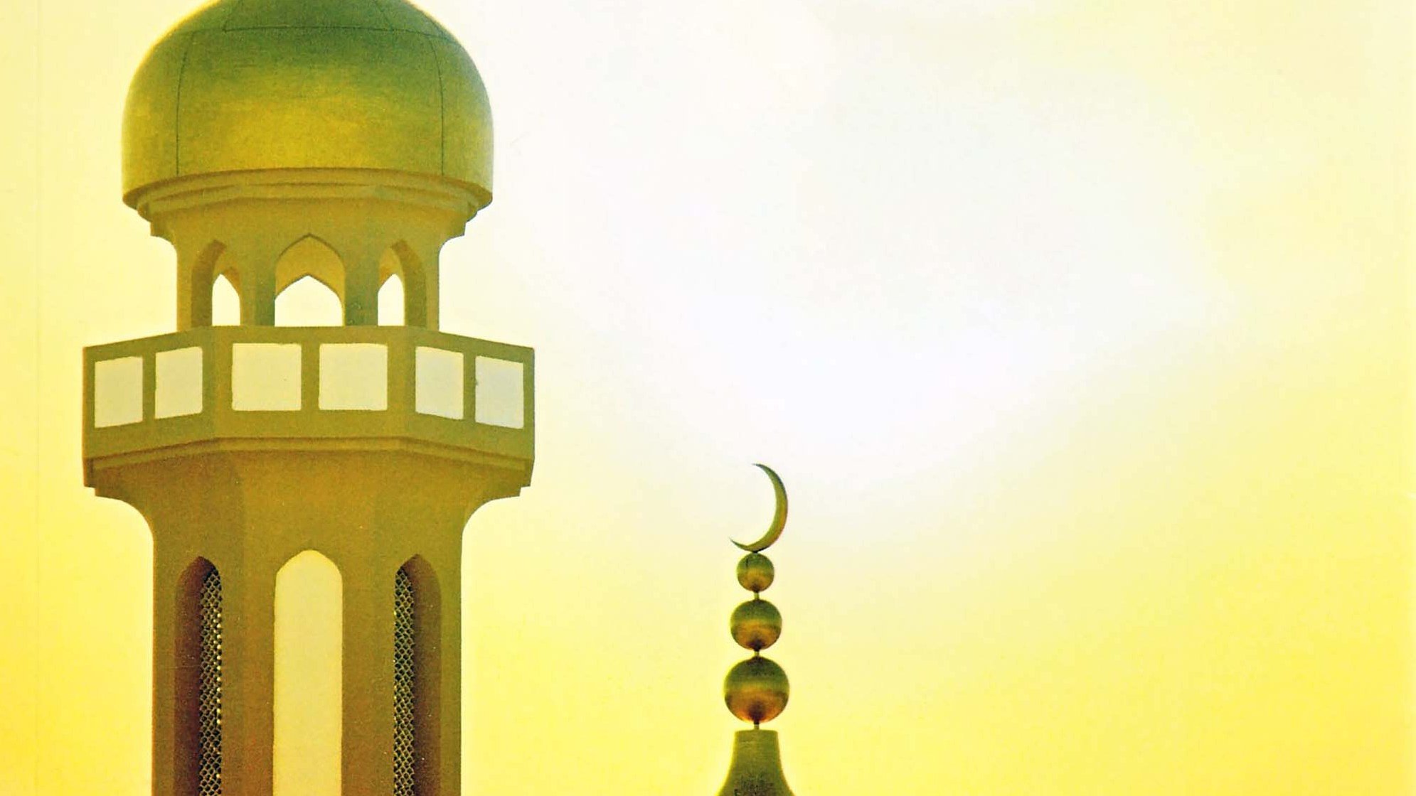 L'immagine mostra il profilo di una moschea, con una cupola grande e una torre alta. La cupola è di forma arrotondata, mentre la torre ha una base decorata con finestre e una punta sormontata da una luna crescente. Sullo sfondo, si intravede un cielo luminoso con tonalità calde di giallo, creando un'atmosfera suggestiva e serena.