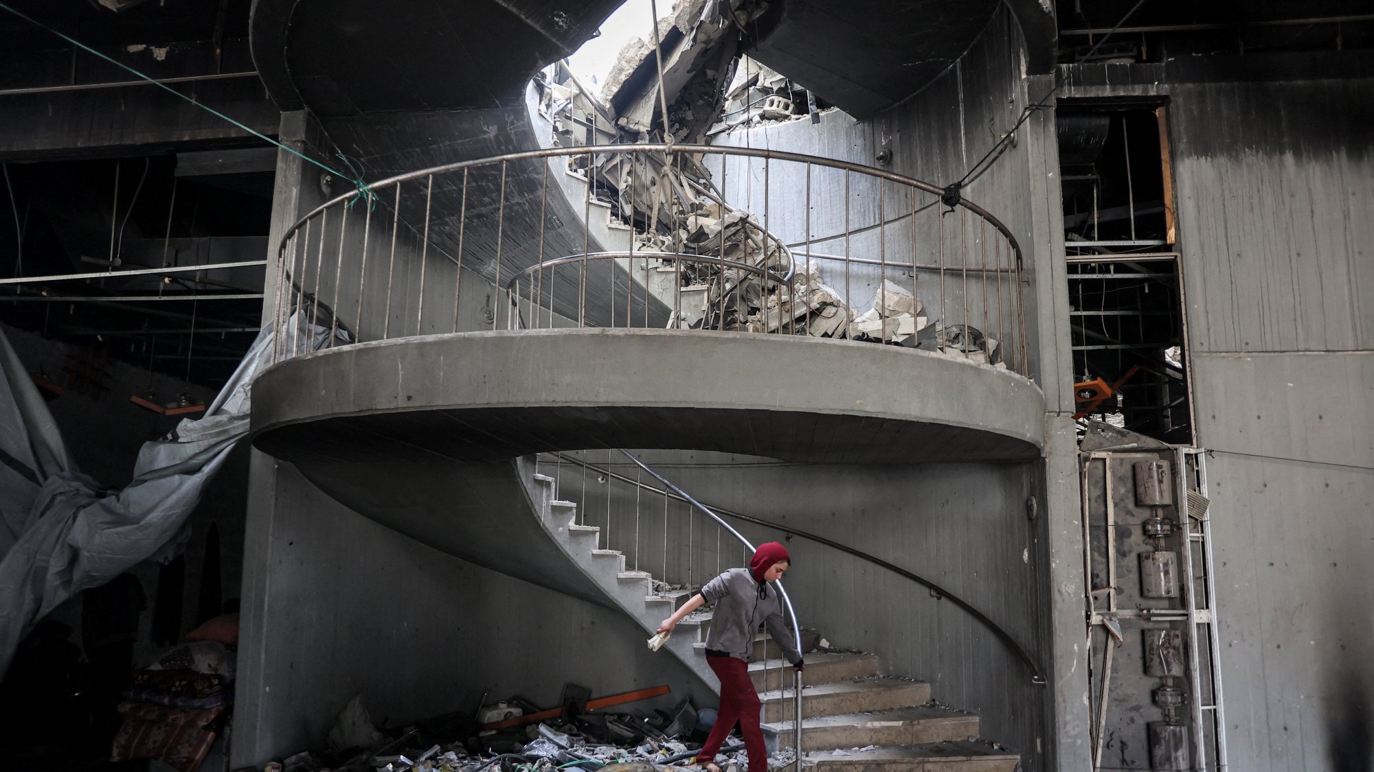 L'immagine mostra una scala a chiocciola danneggiata all'interno di un edificio in rovina. La struttura presenta macerie sparse e parte del soffitto è crollato, creando un'apertura sopra la scala. Una persona, vestita con un abbigliamento rosso e coperta da un copricapo, sta camminando sulla scala tra le macerie. L'atmosfera è quella di un luogo in crisi, con segni evidenti di distruzione e abbandono.