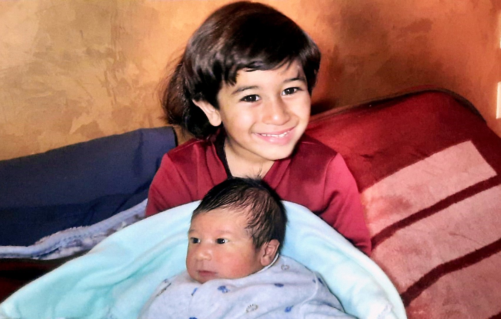 Nell'immagine si vede un bambino più grande che sorride mentre tiene in braccio un neonato avvolto in una coperta blu. Il bambino più grande ha i capelli scuri e lunghi, e indossa una maglietta rossa. Il neonato ha i capelli scuri e un'espressione curiosa. Sullo sfondo ci sono delle coperte sul lettino, creando un'atmosfera accogliente e familiare.