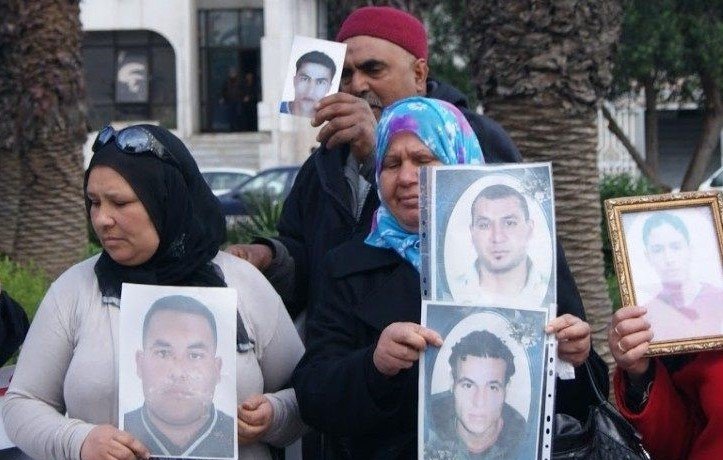 Nell'immagine si vede un gruppo di persone che partecipano a una manifestazione. Alcuni di loro tengono in mano delle fotografie di uomini. Le donne indossano il velo e mostrano espressioni serie, mentre gli uomini dietro di loro sembrano sostenere le stesse fotografie. L'ambiente intorno è urbano, con alberi e edifici visibili sullo sfondo. La scena trasmette un senso di protesta e richiesta di giustizia.