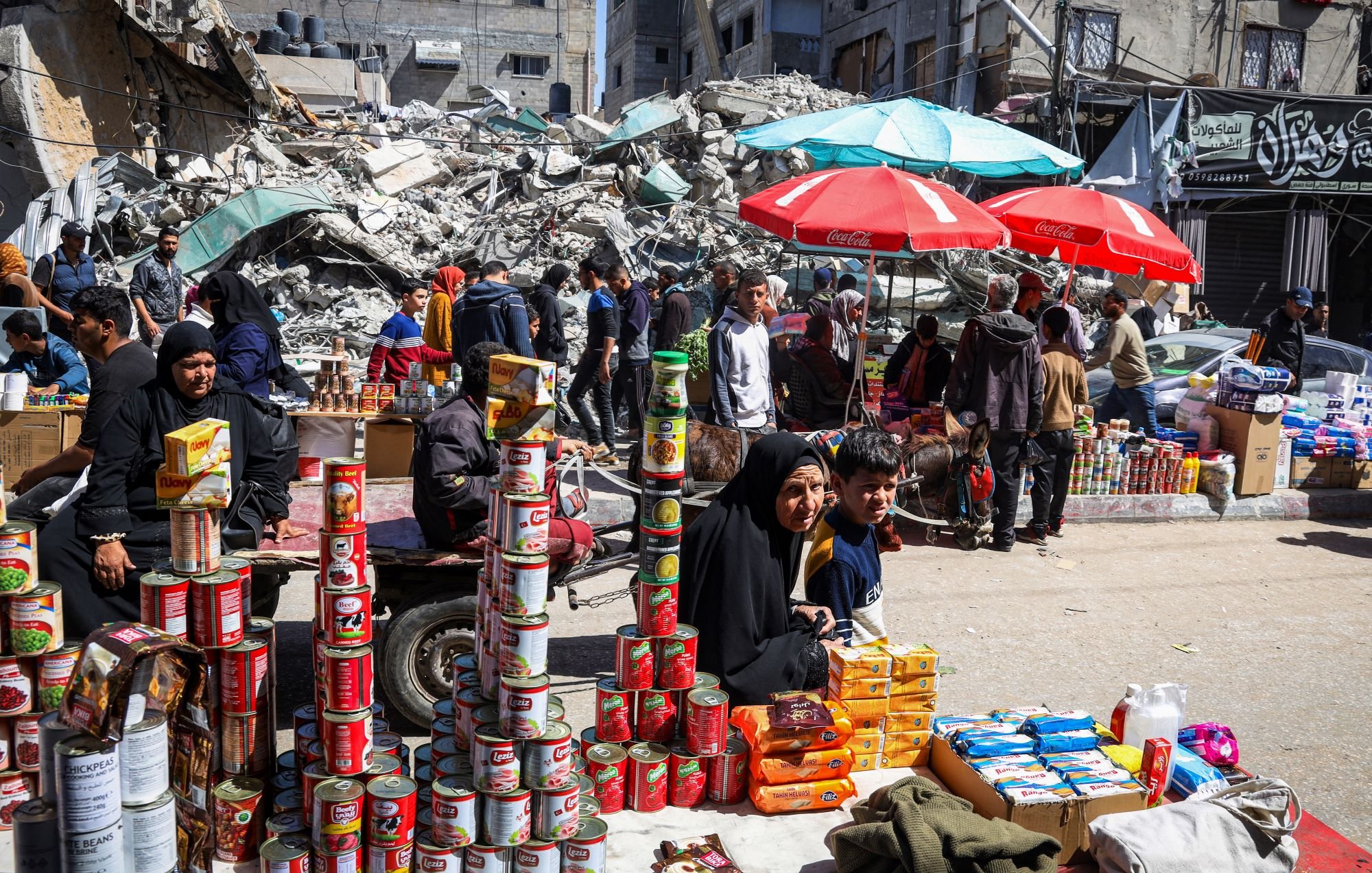 L'immagine mostra una scena di un mercato affollato, situato tra rovine di edifici. In primo piano, un gruppo di persone sta vendendo prodotti alimentari, con diversi ripiani di scatole e cibo in mostra. Sono visibili delle donne e dei bambini, alcuni seduti e altri in piedi, mentre diversi ombrelloni rossi forniscono ombra. Sullo sfondo, ci sono detriti di edifici crollati, che testimoniano una situazione di crisi. L'atmosfera sembra vivace nonostante le difficoltà evidenti.