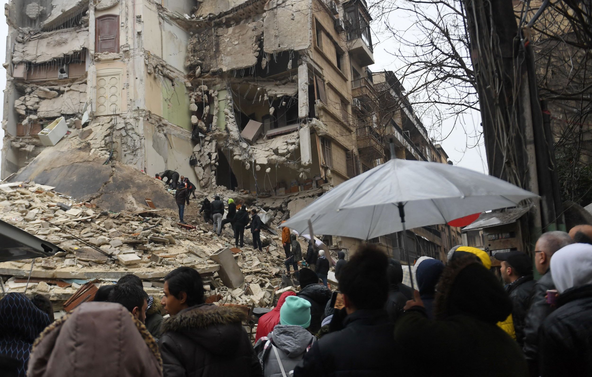 L'immagine mostra una scena dopo un terremoto devastante. Si può vedere un edificio gravemente danneggiato, con pareti crollate e macerie sparse. Alcuni soccorritori stanno cercando di recuperare persone intrappolate tra i detriti. Una folla di persone osserva da una certa distanza, alcune delle quali sono riparate sotto ombrelli, probabilmente a causa della pioggia. L'atmosfera è caratterizzata da tensione e preoccupazione, mentre le squadre di soccorso lavorano per salvare vite.