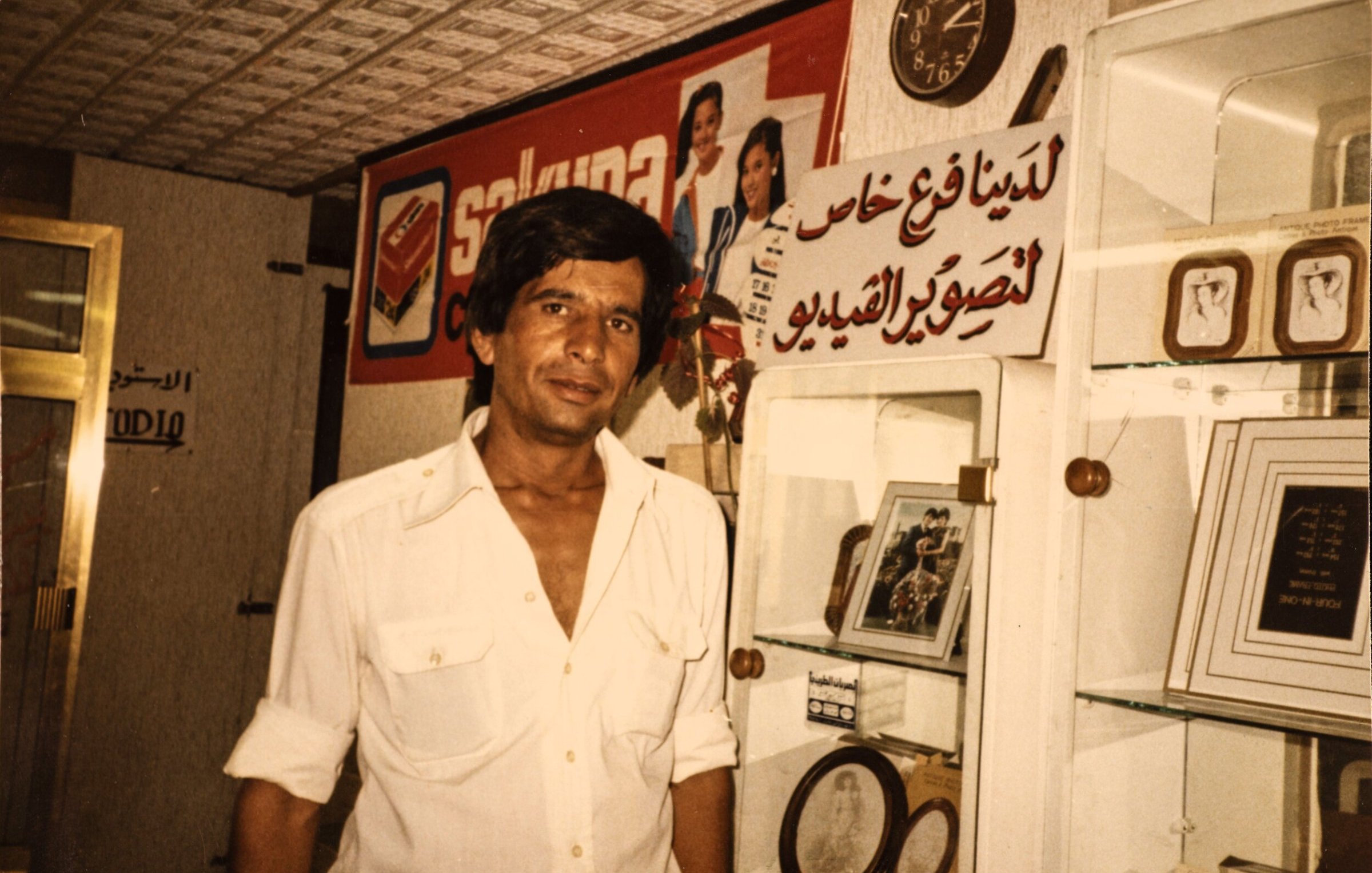 L'immagine mostra un uomo in un ambiente che sembra essere un negozio o un'esposizione. Indossa una camicia bianca e ha i capelli scuri e lunghi. Sullo sfondo, si possono vedere vari oggetti esposti, tra cui quadri e cornici. C'è anche un grande cartello con del testo in arabo e un orologio visibile. L'atmosfera complessiva trasmette un senso di nostalgia.