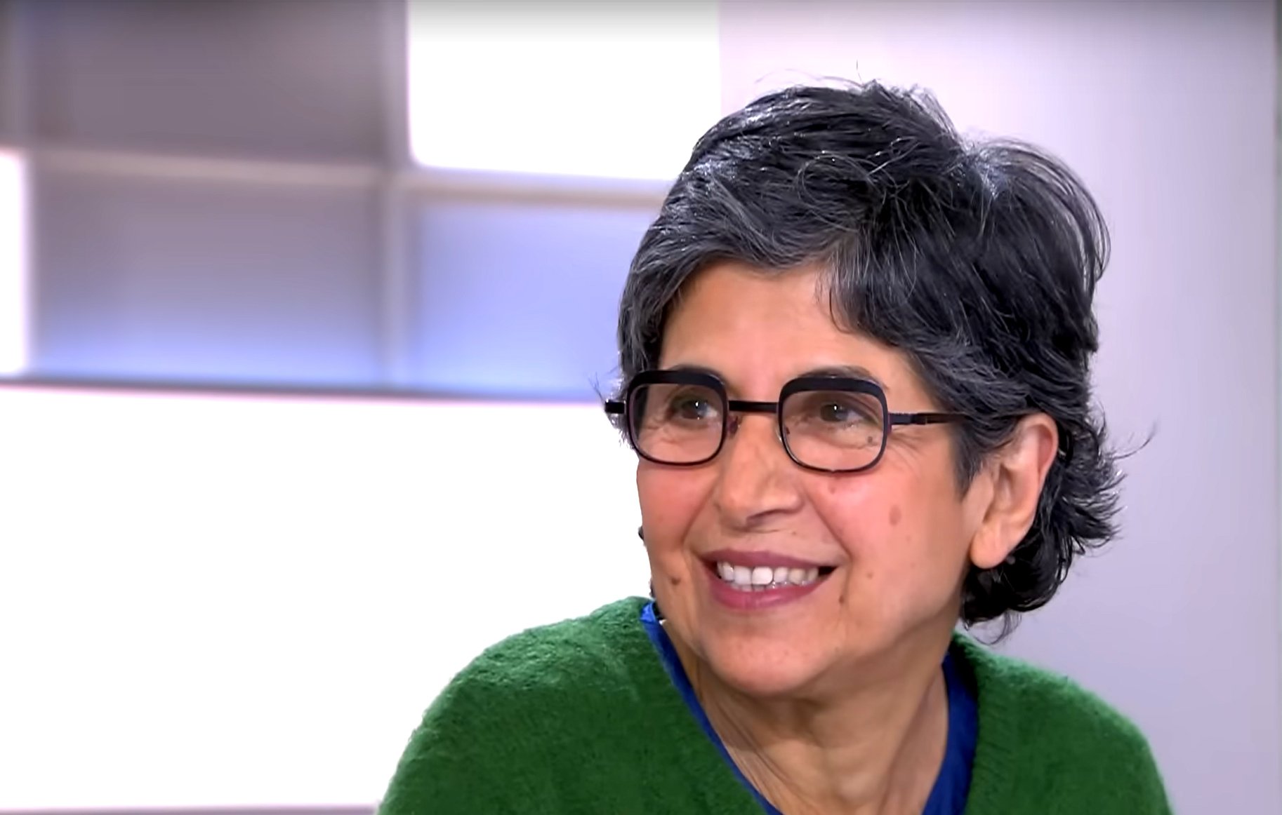 L'immagine mostra una donna con capelli grigi e ondulati, indossando occhiali e un maglione verde. Sembra sorridere e avere un'espressione accogliente. Sullo sfondo ci sono sfumature chiare che danno un'atmosfera luminosa e positiva.