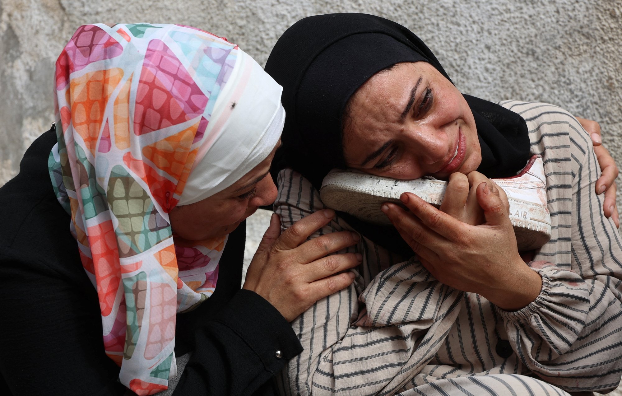 Due donne in abiti tradizionali si abbracciano, una piange e tiene un oggetto vicino al viso.