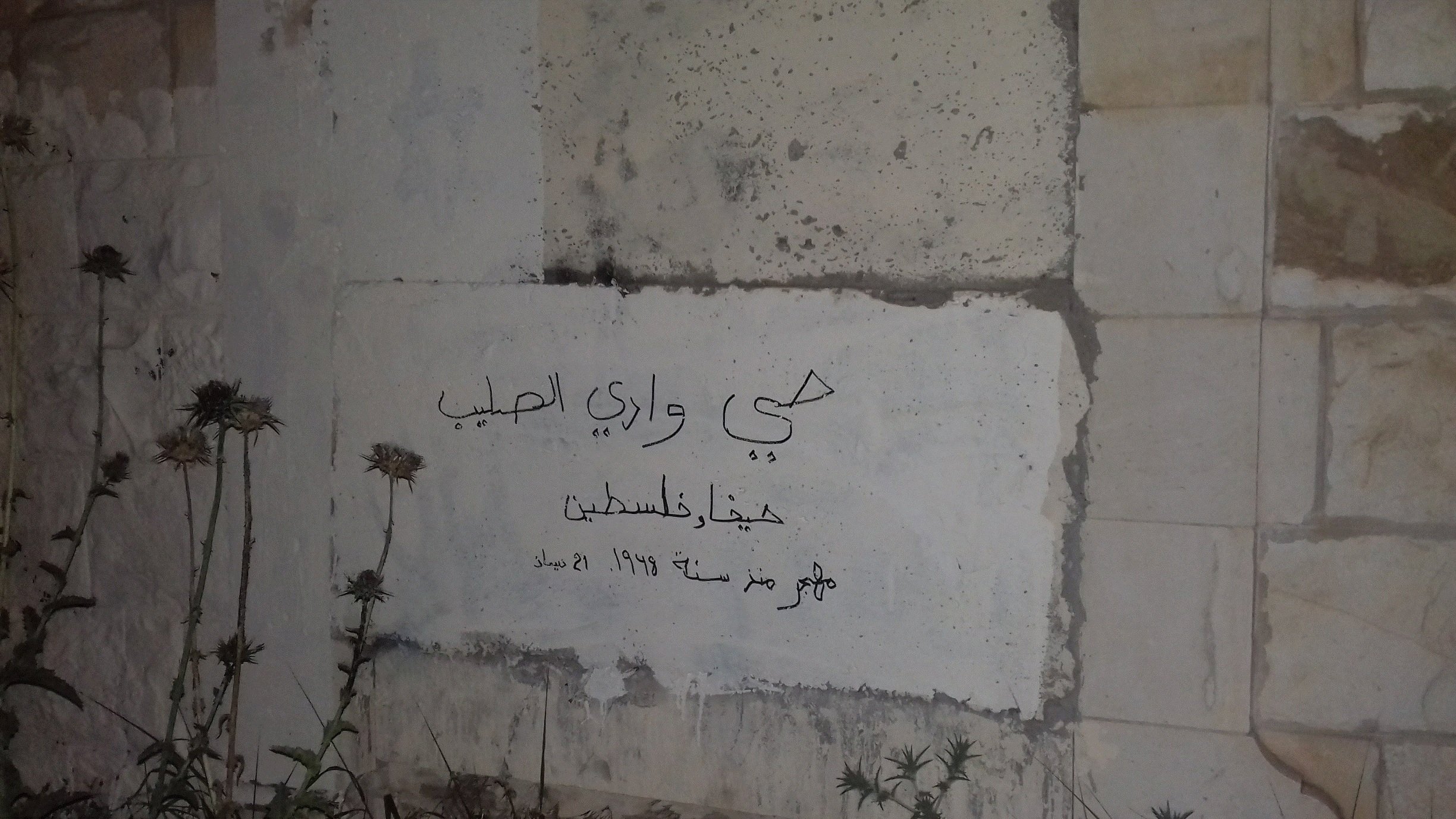 L'immagine mostra una parete di pietra con un'iscrizione in arabo. La scritta fa riferimento a un luogo specifico, con delle date evidenti, probabilmente legate alla storia palestinese. Attorno alla scritta si notano delle erbacce, che suggeriscono che il posto non sia in uso o mantenga un aspetto trascurato. La scena evoca un senso di abbandono e memoria.
