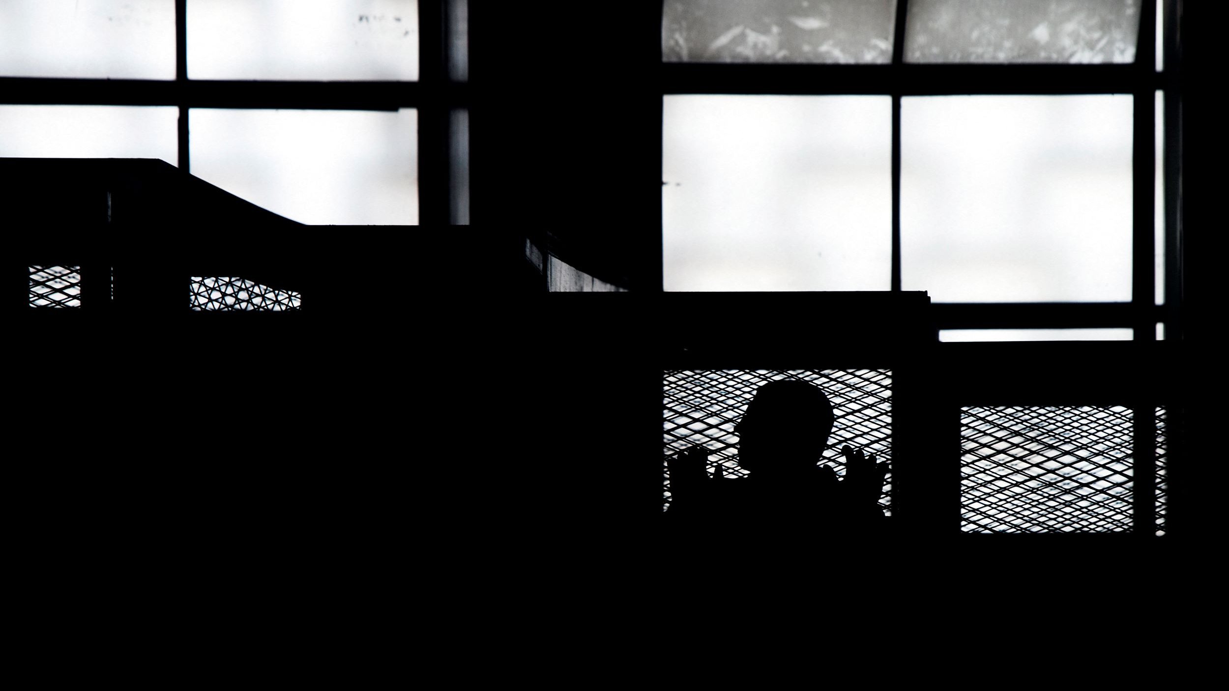 L'immagine presenta una scena in silouette, in cui si intravede la figura di una persona dietro una rete metallica, all'interno di un ambiente che sembra essere un edificio. Le finestre nel fondo lasciano filtrare una luce diffusa, creando un contrasto marcato tra le ombre e la luminosità dello sfondo. L'atmosfera generale risulta suggestiva e misteriosa, enfatizzando la separazione tra la figura e l'ambiente circostante.