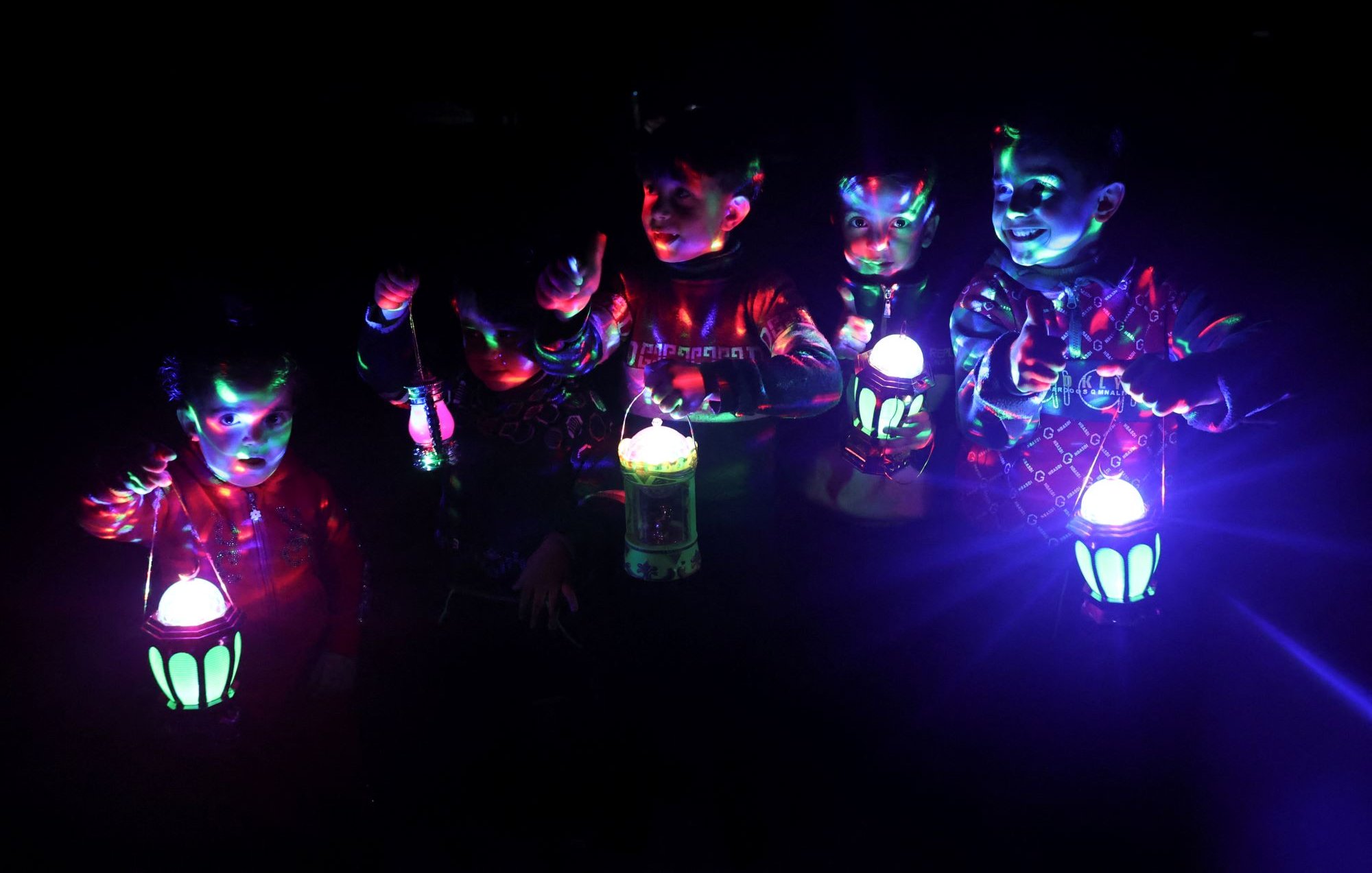 Nell'immagine si possono vedere sei bambini che si trovano in un ambiente buio, illuminati da luci colorate che proiettano effetti dinamici. Ognuno dei bambini tiene una lanterna che emette una luce vivace e colorata. I piccoli sembrano divertirsi, sorridendo e interagendo tra di loro. Le loro espressioni trasmettono gioia e entusiasmo, creando un'atmosfera festosa. Le luci colorate e le ombre contribuiscono a rendere l'immagine molto suggestiva.