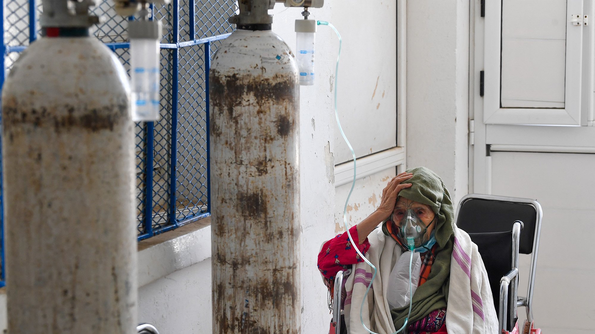 L'immagine mostra una donna anziana seduta su una sedia a rotelle in un ambiente ospedaliero. Indossa una maschera per l'ossigeno e sembra preoccupata, con una mano sulla fronte. Dietro di lei ci sono bombole di ossigeno e un ambiente che appare abbastanza semplice e clinico. L'atmosfera trasmette una sensazione di vulnerabilità e dolore, evidenziando le difficoltà legate alla salute.
