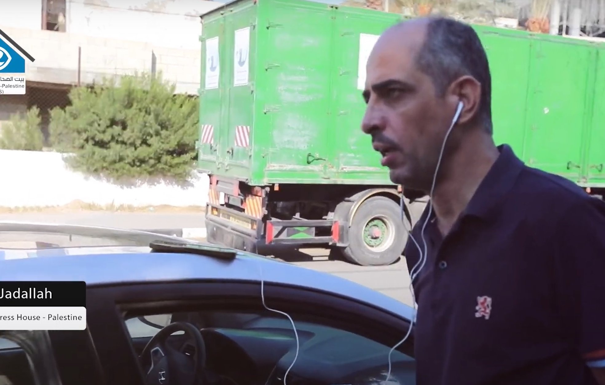 Nell'immagine si vede un uomo che cammina accanto a un'auto. Indossa una maglietta scura e ha delle cuffie che gli pendono dalle orecchie. Sullo sfondo si nota un camion verde e alcune piante. L'uomo sembra concentrato e diretto verso un punto specifico. A sinistra, è presente un'etichetta che indica il suo nome e la sua posizione, come direttore di un'organizzazione legata alla stampa in Palestina.