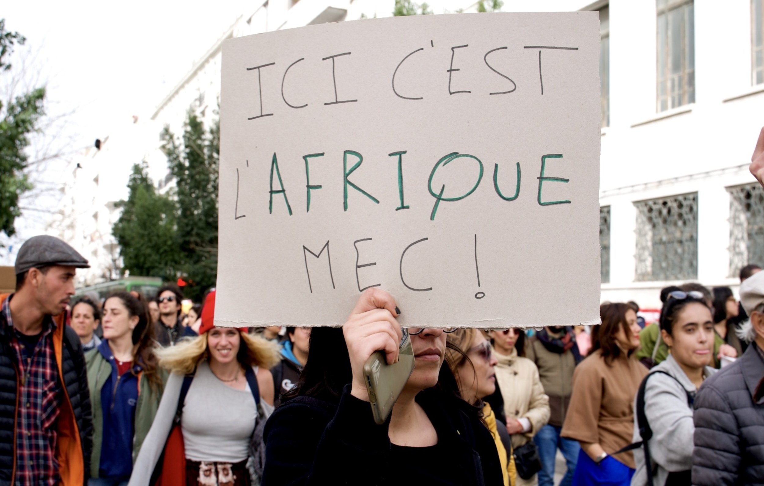 Nell'immagine si vede un gruppo di persone durante una manifestazione. Una donna in primo piano tiene in mano un cartello scritto in francese che recita "ICI C'EST L'AFRIQUE MEC!", il quale esprime un messaggio forte e chiaro. L'atmosfera sembra vivace e partecipativa, con diversi partecipanti che si uniscono per sostenere una causa. Sullo sfondo si notano alberi e edifici, suggerendo un contesto urbano.