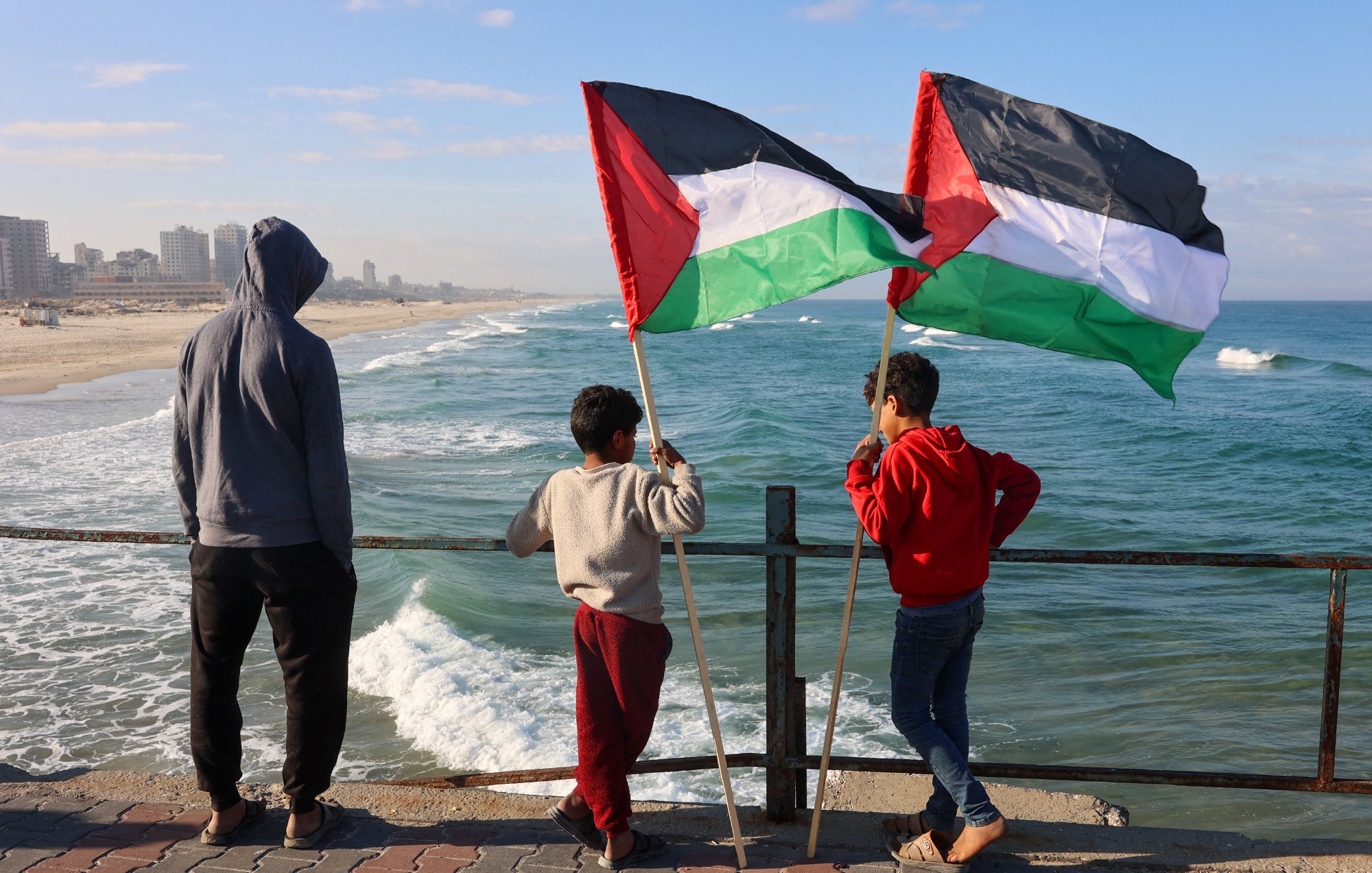 Nell'immagine, tre ragazzi sono in piedi su un molo che si affaccia su una spiaggia e sul mare. Due di loro tengono in alto delle bandiere palestinesi, che sono di colore nero, bianco, verde e rosso. Il ragazzo più piccolo, in una felpa grigia, sembra osservare attentamente l'orizzonte. La scena è illuminata da una luce naturale, con il cielo blu e alcune nuvole sparse. Le onde del mare si infrangono dolcemente sulla riva, creando un'atmosfera tranquilla. Gli altri due ragazzi indossano abbigliamento casual e sembrano condividere un momento di riflessione insieme.