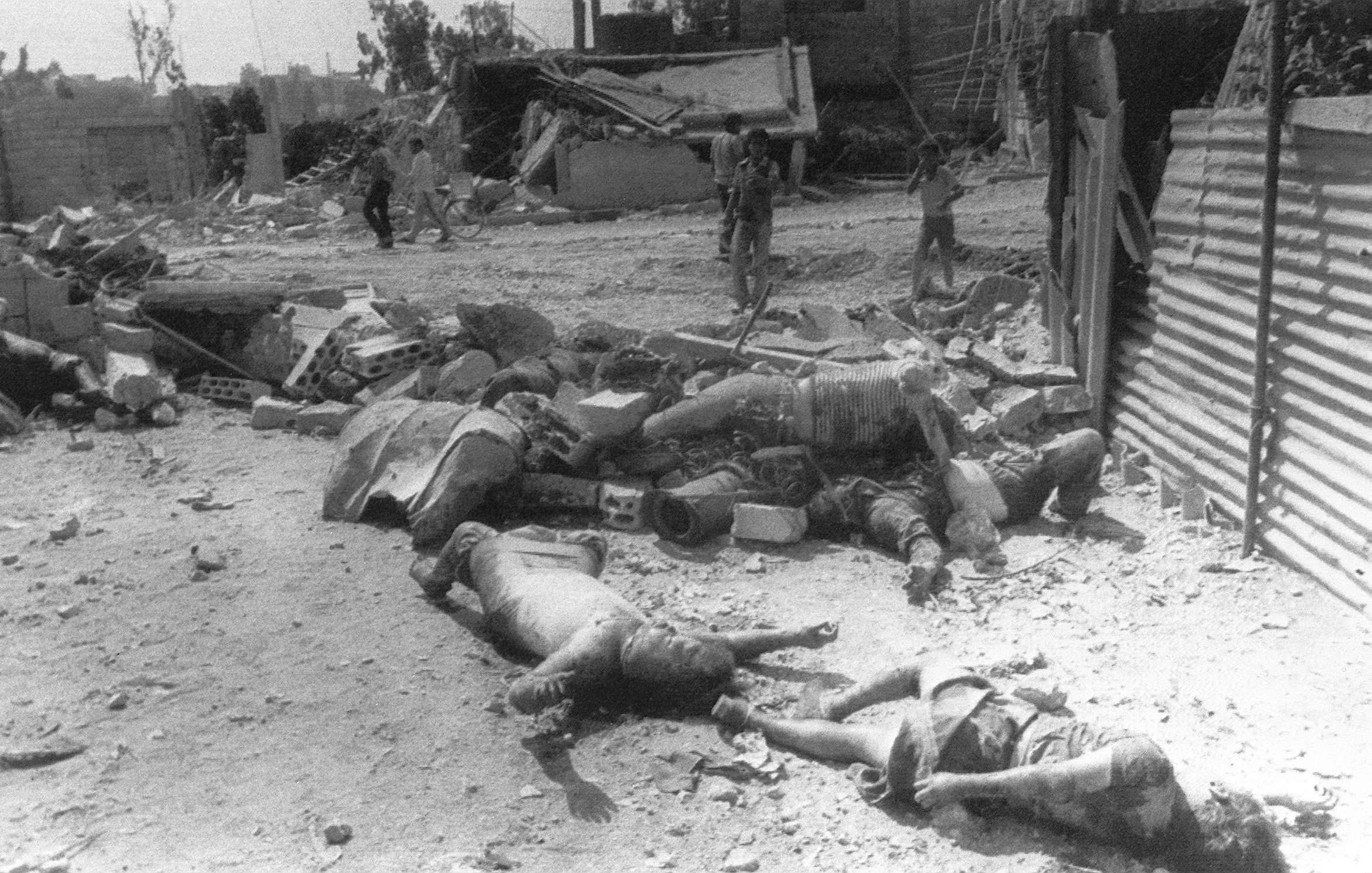 L'immagine mostra un ambiente devastato, probabilmente dopo un attacco o un evento distruttivo. Sul terreno ci sono corpi senza vita, apparentemente tra le macerie di edifici distrutti. In lontananza, alcune figure umane sembrano muoversi tra i detriti. La scena evoca un forte senso di sofferenza e desolazione, riflettendo gli effetti devastanti della violenza o di un conflitto.