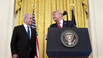 L'immagine mostra due uomini in un ambiente ufficiale, probabilmente in una cerimonia o conferenza stampa. Sono di fronte a un podio con il sigillo presidenziale degli Stati Uniti. Sullo sfondo, ci sono delle tende dorate e le bandiere statunitensi. I due uomini sembrano sorridere e interagire in modo cordiale.