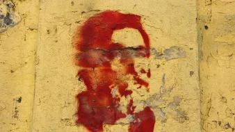 L'immagine mostra un murale su una parete di colore giallo. Il murale rappresenta un volto stilizzato, probabilmente un personaggio noto, dipinto in rosso. Sotto il volto ci sono delle scritte, anch'esse in rosso, che sembrano in arabo. La superficie della parete appare consumata e scrostata, contribuendo a un aspetto storico e di protesta.