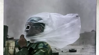 L'immagine mostra un uomo in uniforme militare, avvolto da un velo di stoffa che sembra fluttuare nel vento. Il suo viso è parzialmente coperto, rendendolo quasi irriconoscibile. Sullo sfondo si intravede un paesaggio desolato, con sabbia e detriti, suggerendo che si trovi in un ambiente arido, probabilmente in una zona di conflitto. L'atmosfera appare drammatica a causa del vento che solleva la polvere.