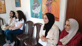 Nell'immagine si vedono quattro donne sedute su delle sedie in una stanza. Le pareti sono decorate con opere d'arte colorate, rappresentanti differenti stili. Le donne indossano abbigliamenti diversi: alcune hanno il velo, altre hanno abiti casual. Il loro atteggiamento sembra tranquillo, mentre osservano l'ambiente circostante. La luce che entra dalla finestra illumina la scena, creando un'atmosfera accogliente.