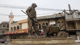 L'immagine mostra una statua di un operaio che lavora, con una pala in mano e una carriola accanto a lui. Il monumento è situato in una zona urbana, caratterizzata da edifici danneggiati e in rovina sullo sfondo. La scena trasmette un senso di resilienza e dignità, riflettendo la vita e il lavoro in un contesto difficile. L'atmosfera è un po' desolata a causa delle strutture danneggiate, mentre la statua rappresenta il duro lavoro e l'impegno quotidiano delle persone.