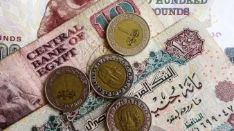 L'immagine mostra diverse banconote e monete egiziane. In primo piano ci sono quattro monete, due delle quali sono di un pound egiziano. Sullo sfondo, ci sono banconote di diverse denominazioni, evidentemente con scritte arabe, tipiche della valuta egiziana. Una banconota sembra avere il valore di cento pounds e presenta un motivo decorativo. L'atmosfera è quella di un contesto economico, evidenziando elementi tipici della valuta dell'Egitto.