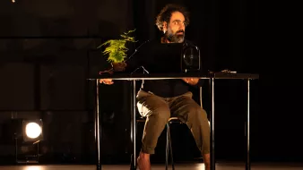 L'immagine mostra un uomo seduto a un tavolo in un ambiente di scena illuminato. Indossa una maglietta nera e pantaloni chiari, e sembra concentrato su un oggetto davanti a lui. Accanto a lui c'è una piccola pianta in vaso, mentre una ventola è posizionata sulla sua sinistra. L'illuminazione crea un'atmosfera drammatica, evidenziando sia l'uomo che gli oggetti sul tavolo. L'ambiente è scuro, con alcune luci che si riflettono sul pavimento.