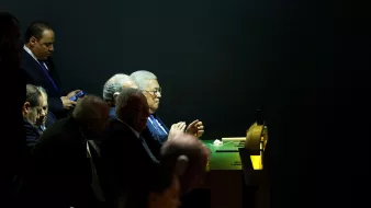 L'immagine mostra un gruppo di uomini seduti a un tavolo, apparentemente impegnati in una discussione o in un evento ufficiale. Alcuni di loro sembrano concentrati, mentre uno di loro sta guardando il cellulare. L'illuminazione è tenue, con un contrasto tra le zone illuminate e quelle in ombra, conferendo un'atmosfera seria e formale all'ambiente. Non ci sono elementi di identificazione chiara per i volti dei partecipanti.