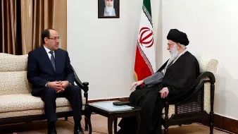 Due uomini seduti in una sala con bandiera iraniana, discutono seriamente.