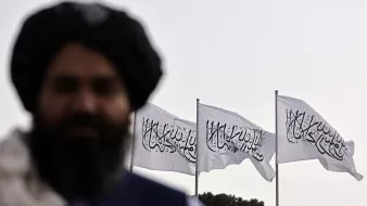 L'immagine mostra un uomo non completamente a fuoco in primo piano, con una folta barba e un turbante. Sullo sfondo, ci sono tre bandiere bianche che sventolano, ognuna recante un simbolo o una scritta. Le bandiere evocano un contesto culturale e politico specifico, tipico di alcune regioni. L'atmosfera è suggestiva e mette in risalto il contrasto tra il soggetto in primo piano e lo sfondo.