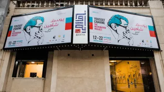 L'immagine mostra un edificio con una facciata decorata da grandi manifesti. I manifesti annunciano un progetto artistico intitolato "The Restoration Project" di Youssef Chahine, con date di evento indicate dal 12 al 22 settembre. Si può notare un design grafico accattivante, con illustrazioni e testo in arabo e in inglese. La zona sembra essere un luogo di cultura o un centro espositivo, con elementi architettonici tipici di un contesto urbano.