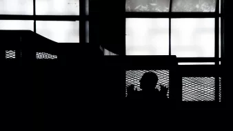 L'immagine presenta una scena in silouette, in cui si intravede la figura di una persona dietro una rete metallica, all'interno di un ambiente che sembra essere un edificio. Le finestre nel fondo lasciano filtrare una luce diffusa, creando un contrasto marcato tra le ombre e la luminosità dello sfondo. L'atmosfera generale risulta suggestiva e misteriosa, enfatizzando la separazione tra la figura e l'ambiente circostante.