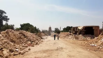 L'immagine mostra una strada sterrata, circondata da macerie e detriti. Sullo sfondo si intravede un edificio con una cupola, probabilmente una moschea o un monumento, avvolto da polvere. La scena trasmette un senso di devastazione, con alberi e strutture danneggiate ai lati della strada. Alcune persone sono visibili in lontananza, mentre il cielo appare chiaro e luminoso.