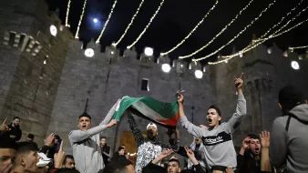 L'immagine mostra un gruppo di persone riunite in un contesto festivo, probabilmente in un evento pubblico. I partecipanti stanno sventolando una bandiera, che sembra essere quella palestinese, e sono visibilmente entusiasti. Sullo sfondo, si può notare una struttura medievale, che suggerisce un'atmosfera di celebrazione. Le luci appese creano un'atmosfera suggestiva e notturna, mentre i volti delle persone esprimono gioia e partecipazione.