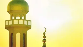 L'immagine mostra il profilo di una moschea, con una cupola grande e una torre alta. La cupola è di forma arrotondata, mentre la torre ha una base decorata con finestre e una punta sormontata da una luna crescente. Sullo sfondo, si intravede un cielo luminoso con tonalità calde di giallo, creando un'atmosfera suggestiva e serena.