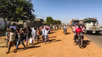 L'immagine mostra una scena di vita quotidiana lungo una strada in un ambiente urbano o semi-urbano. Si notano molte persone che camminano lungo il lato della strada, alcune con sacchi o beni in mano, e altre che sembrano discutere tra di loro. Sul lato opposto della strada ci sono veicoli in transito, tra cui camion e motociclette. La vegetazione circostante è presente, con alcuni alberi che offrono ombra. Il terreno appare polveroso, e l'atmosfera complessiva sembra vivace e movimentata.