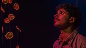 L'immagine mostra un giovane uomo che osserva con meraviglia degli agrumi essiccati appesi in aria. La sua espressione è attenta e sognante, mentre le luci soffuse creano un'atmosfera suggestiva. I colori caldi degli agrumi contrastano con il fondo scuro, enfatizzando la bellezza dell'installazione.