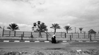 L'immagine ritrae una scena in bianco e nero che mostra una figura solitaria che cammina lungo una strada. Sullo sfondo, si possono vedere alcune palme e una ruota panoramica, che suggerisce la presenza di un parco o di un'area ricreativa. La composizione offre un'atmosfera malinconica e riflessiva, accentuata dalla scelta dei toni scuri e dalla mancanza di colori vivaci. La figura, vestita con abiti tradizionali, sembra essere in un momento di solitudine all'interno di un paesaggio urbano.