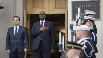 L'immagine mostra due uomini in piedi davanti all'entrata di un edificio governativo, probabilmente il Dipartimento della Difesa. Uno di loro indossa un abbigliamento formale con un completo scuro e una cravatta, mentre l'altro ha il braccio destro sul cuore, indicando un gesto di rispetto o di onore. Dietro di loro, ci sono diversi membri delle forze armate in uniforme, schierati con armi in mano, pronti a rendere onore. L'atmosfera è solenne e pomposa, suggerendo un evento ufficiale o una cerimonia.