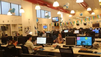 L'immagine mostra un ambiente di lavoro in un ufficio moderno. Si possono vedere diverse scrivanie con computer e monitor. Alcune persone sono immerse nel loro lavoro, mentre altre interagiscono tra di loro. L'ufficio è decorato con palloncini blu e festoni colorati, creando un'atmosfera vivace e informale. Le pareti sono adornate con fotografie e materiali di lavoro, e la luce naturale entra attraverso le grandi finestre.