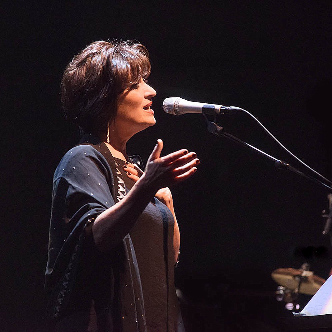 الصورة تظهر سيدة واقفة أمام ميكروفون، وهي تؤدي أغنية أو تتحدث على المسرح. تبدو مرتدية ملابس أنيقة وتعبّر عن مشاعرها من خلال حركات يديها. الإضاءة خافتة مما يضفي جواً درامياً على المشهد، ويوجد في الخلفية موسيقيون مجهولون. تعكس الصورة لحظة فنية جميلة.