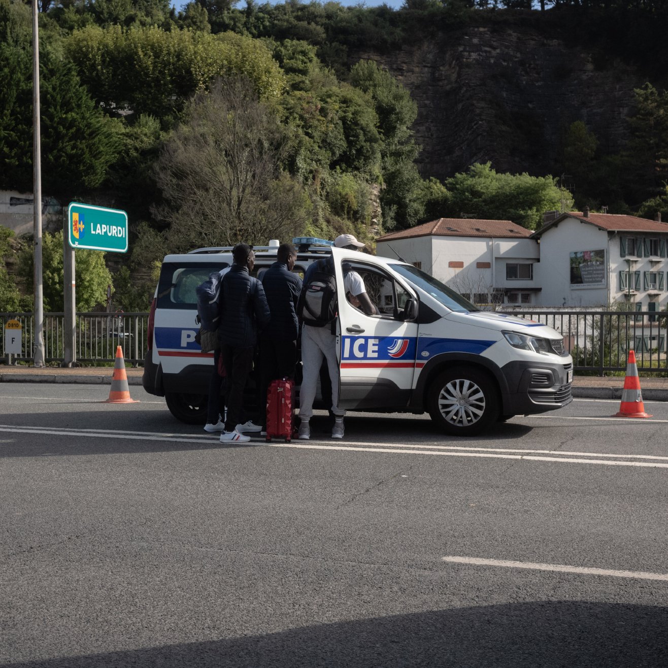The image shows a scene on a road where a police vehicle is stopped. A few individuals are standing near the back of the police car, which is marked with the word "Police." The setting appears to be a highway or a major road, with some road cones placed nearby. In the background, there are trees and some buildings visible, suggesting an urban or suburban area. The atmosphere seems to indicate a police check or an interaction taking place.
