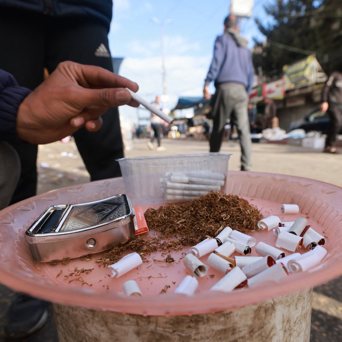L'immagine mostra una persona che tiene in mano una sigaretta su un piatto di plastica rosa, sul quale sono visibili numerosi mozziconi di sigaretta. Accanto ai mozziconi c'è del tabacco sfuso e un contenitore trasparente. Lo sfondo rivela una strada affollata, con persone che si muovono e diversi cartelloni pubblicitari. L'atmosfera sembra quella di un mercato o di una zona urbana.