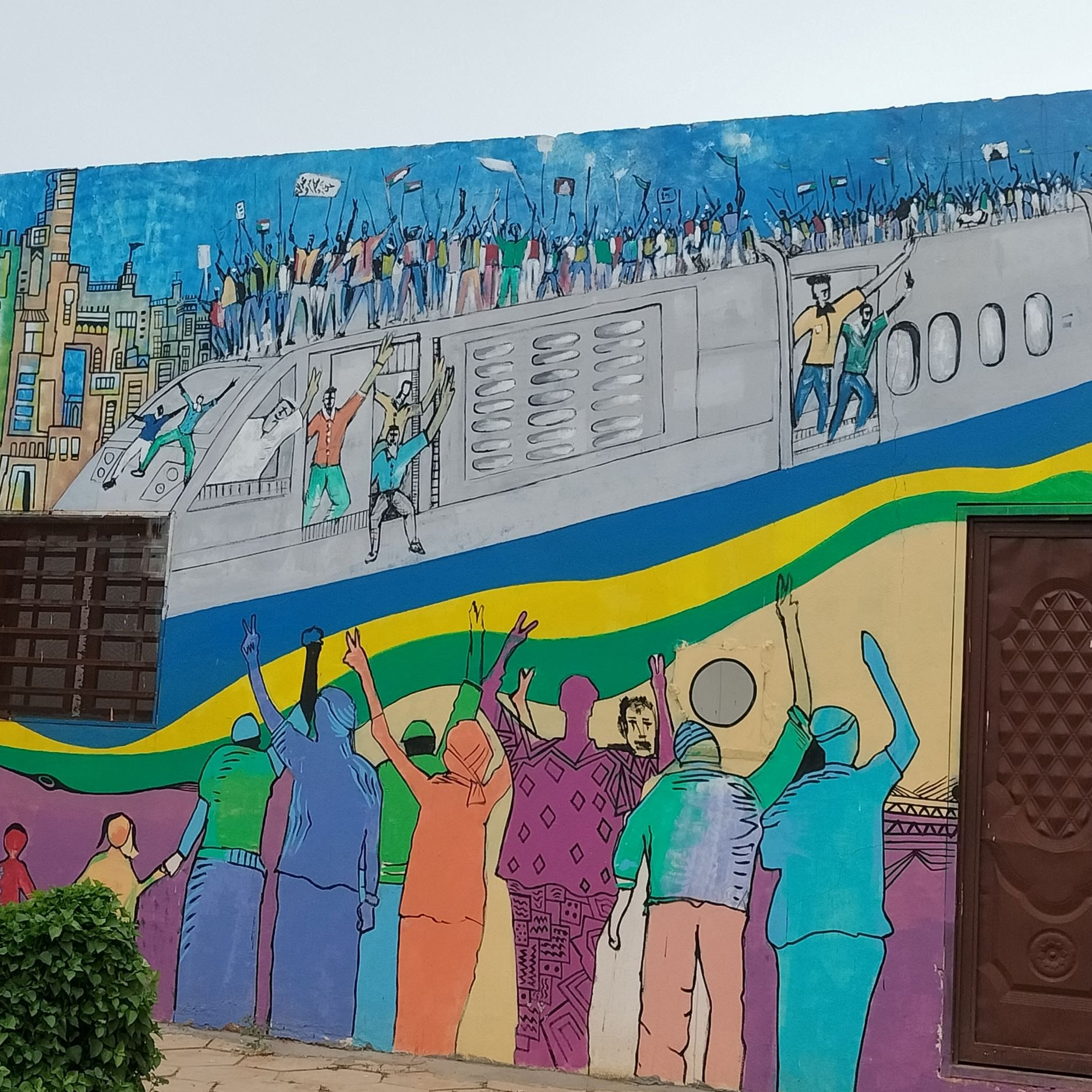 الصورة تظهر جدارية ملونة على حائط، تتضمن مشهدًا حيويًا يبرز مجموعة من الأشخاص الذين يحتفلون أو يتظاهرون. في الجزء العلوي، يظهر مجموعة من الأشخاص على ما يبدو في حالة احتفال، مع أعلام وألوان زاهية. الجزء السفلي يُظهر أشخاصًا يرفعون أيديهم، مما يُعبر عن الحماس أو التأييد. الألوان المستخدمة في الجدارية متنوعة وتجعل الصورة ملفتة للنظر، مما يعطي شعورًا بالحركة والنشاط.