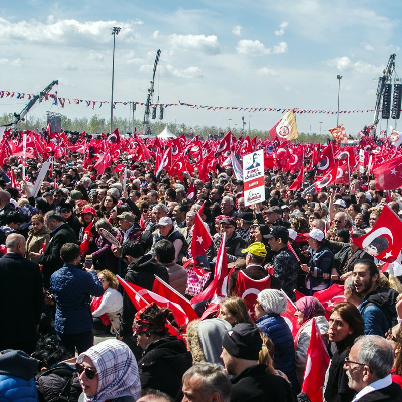 تصویر نشان‌دهنده تظاهرات بزرگی در یک فضای باز است که تعداد زیادی از مردم حضور دارند. آنها پرچم‌های قرمز و سفید ترکیه را در دست دارند و به نظر می‌رسد که در حال ابراز همبستگی یا حمایت از یک حرکت یا رویداد خاص هستند. آسمان آبی است و چند ابر سفید در آن دیده می‌شود. جمعیت متنوعی در تصویر وجود دارد که نشان‌دهنده حضور افراد با سنین و زمینه‌های مختلف اجتماعی است. فعالیت و شور و حال در این جمعیت به وضوح قابل مشاهده است.