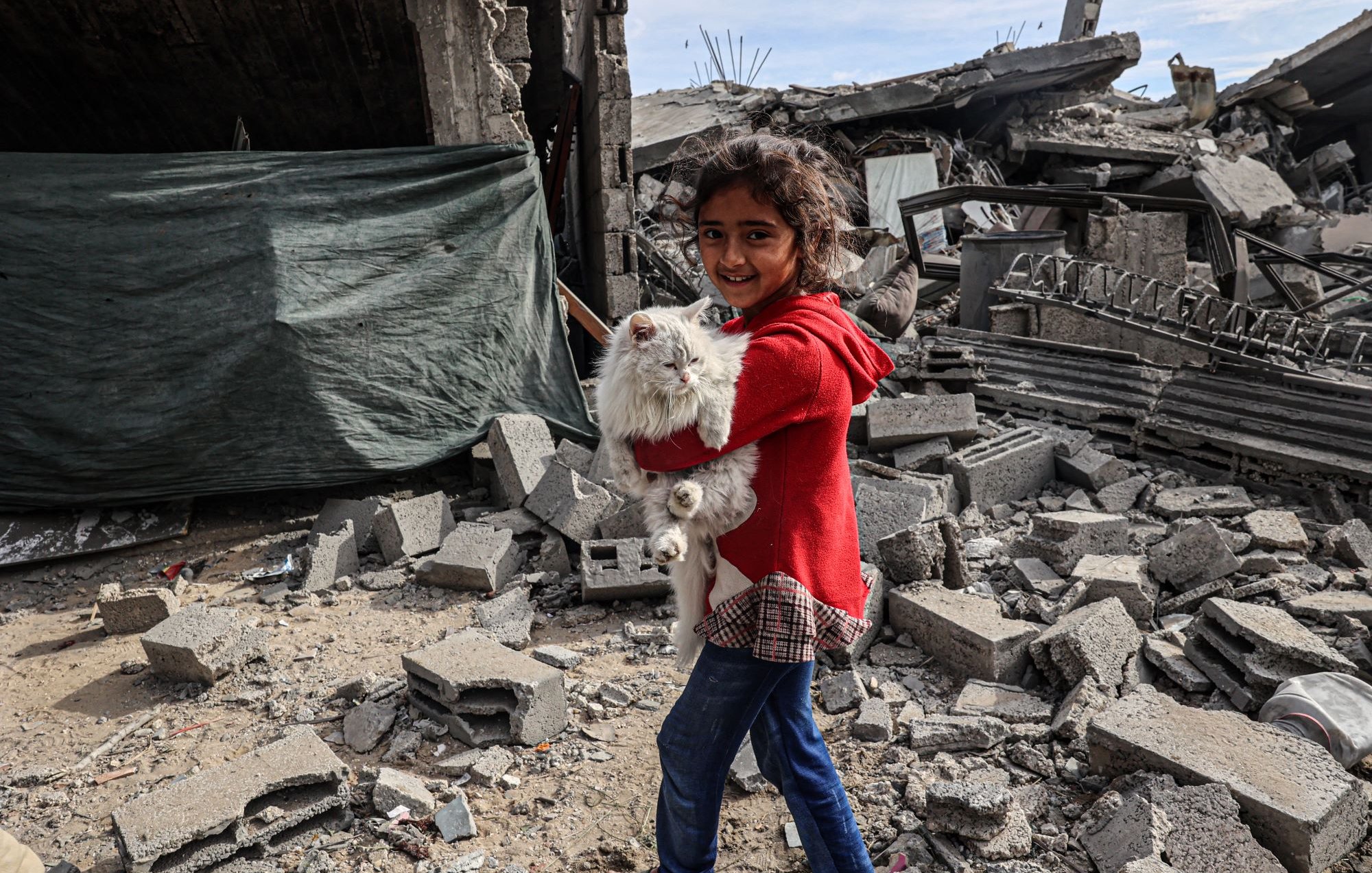 Sur cette image, on voit une jeune fille souriante portant un chat blanc dans ses bras. Elle est habillée d'un manteau rouge et d'un jean bleu. La scène se déroule au milieu de décombres, avec des bâtiments détruits et des débris tout autour. Le ciel est partiellement visible et semble être couvert de nuages. L'image donne une impression de résilience et d'espoir malgré un environnement de destruction.