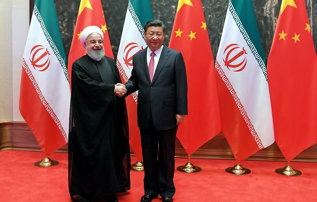 The image depicts two men shaking hands, standing in front of flags of Iran and China. The man on the left is wearing a black robe and white turban, representing Iran, while the man on the right is dressed in a suit. They appear to be in a formal setting, likely symbolizing a diplomatic meeting between the two countries. The background features a red carpet and prominently displayed national flags.