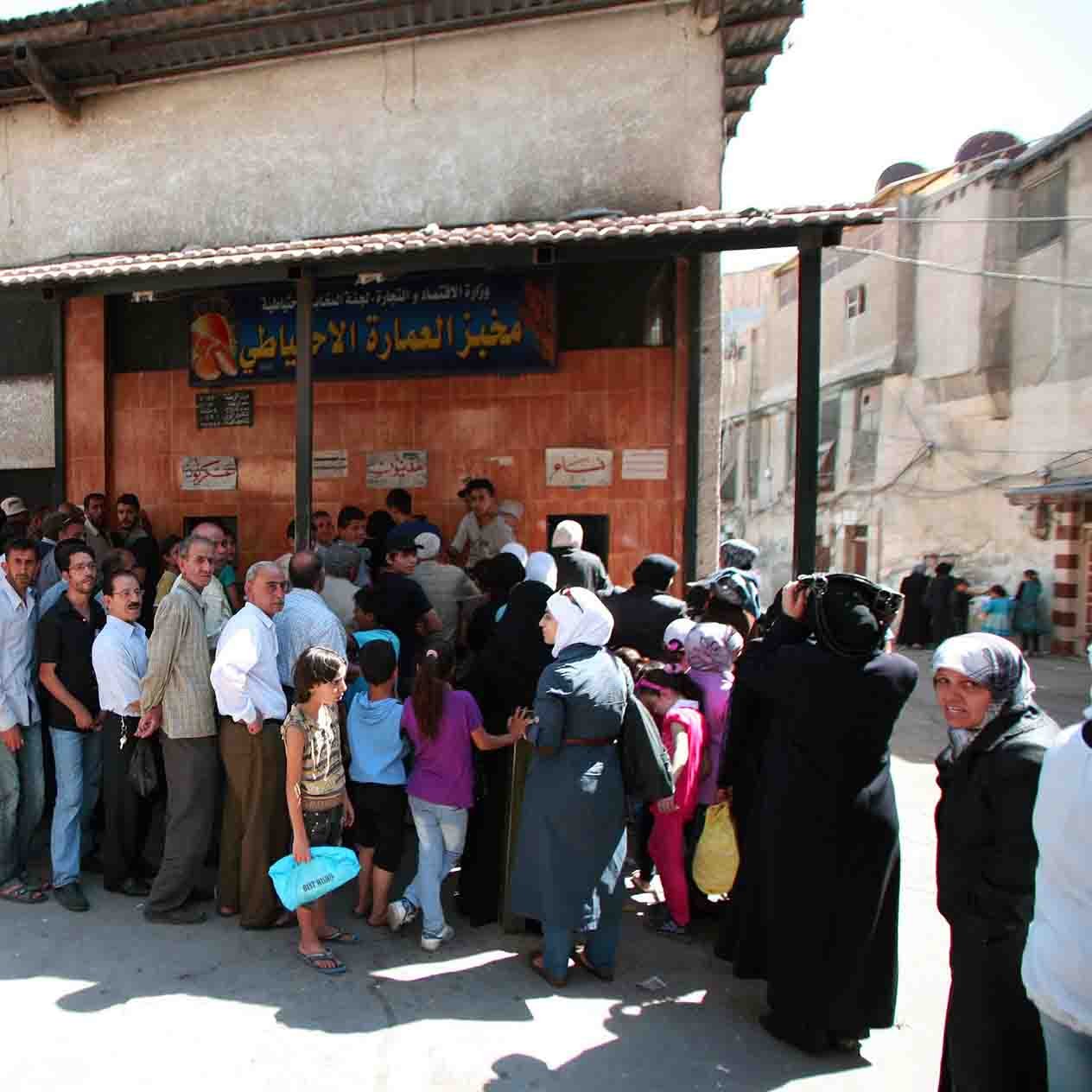 در تصویر، گروهی از مردم به صف ایستاده‌اند. ساختمان در پس‌زمینه دارای نمای ساده‌ای است و تابلو بزرگی بالای درب ورودی نصب شده است. افراد در صف شامل مردان و زنانی هستند که برخی از آن‌ها سبدها یا کیسه‌های خرید را در دست دارند. نور خورشید قوی به تصویر تابیده و فضا شلوغ به نظر می‌رسد. برخی از افراد به طرف دوربین نگاه می‌کنند و برخی دیگر مشغول صحبت هستند. دوچرخه‌ها نیز در کنار خیابان به چشم می‌خورند.