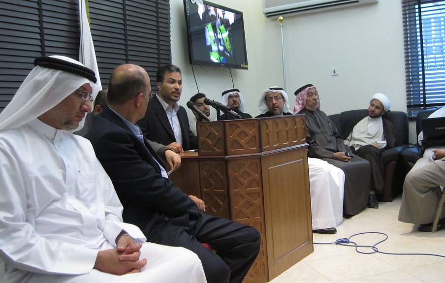 L'image montre un groupe de personnes rassemblées dans une salle. Au centre, un homme est en train de parler devant un microphone, tandis que les autres l'écoutent attentivement. Certains d'entre eux portent des vêtements traditionnels, comme des kandouras et des ghutras, indiquant peut-être un contexte culturel spécifique. À l'arrière-plan, un écran affiche probablement une présentation ou une vidéo. L'atmosphère semble sérieuse et concentrée, suggérant que la réunion pourrait porter sur un sujet important.