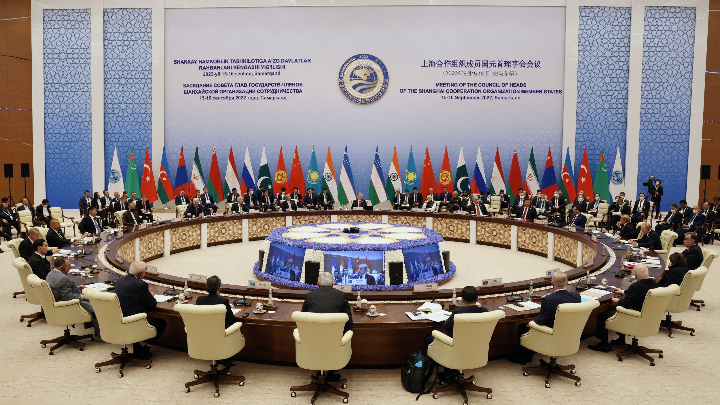 تظهر الصورة اجتماعاً رسمياً حيث يجلس عدد من القادة والممثلين حول طاولة دائرية كبيرة. يتميز المكان بزينة أنيقة، والجدران مزينة بأعلام دول مختلفة. يبدو أن هذا الاجتماع يهدف إلى مناقشة قضايا مهمة، حيث يظهر المشاركون في شكل جاد ومركز. تمثل الطاولة الكبيرة مركز الاجتماع، بينما تتواجد لافتات تشير إلى الحدث والمكان الذي يُعقد فيه الاجتماع.