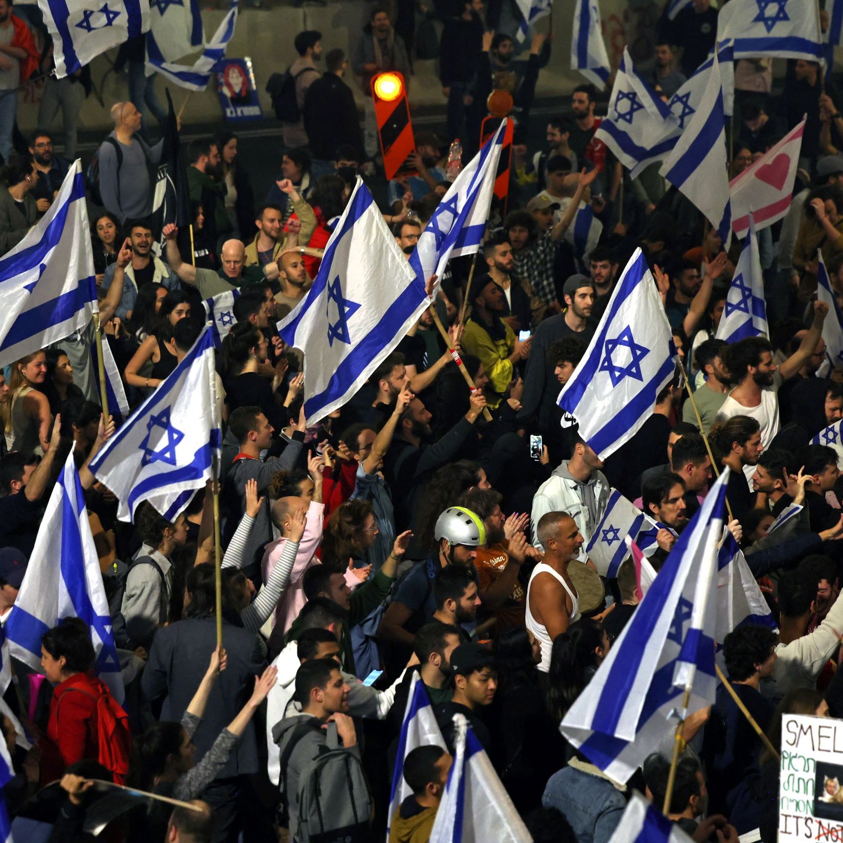 L'immagine mostra una grande folla di persone riunite in un ambiente all'aperto, probabilmente durante una manifestazione. La maggior parte dei partecipanti tiene in mano bandiere di Israele, caratterizzate da una stella di David. Si possono notare anche cartelli, alcuni dei quali contengono frasi e messaggi, mentre altri partecipanti sembrano essere molto energici e coinvolti nell'evento. L'atmosfera appare vibrante e animata, con una forte presenza di colori e simboli nazionali.