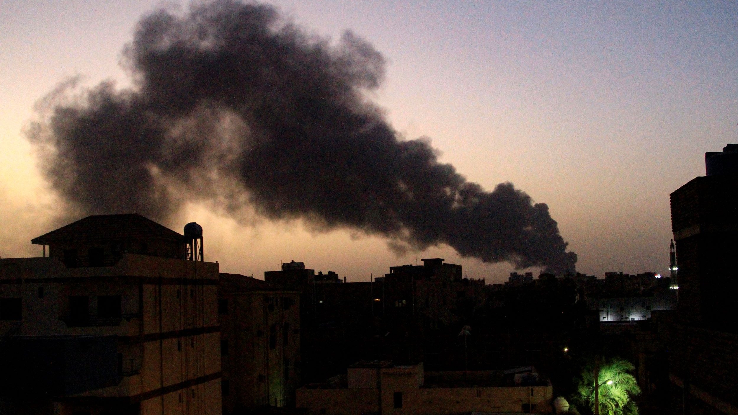 L'immagine mostra un paesaggio urbano al crepuscolo, con un cielo che tende al blu scuro. In lontananza si solleva una colonna di fumo nero, probabilmente proveniente da un incendio o una fonte di combustione. Gli edifici in primo piano sono silhouette scure contro il cielo, e si possono scorgere alcune luci artificiali che brillano tra le strutture. La vegetazione, come le palme, è visibile sul lato destro, aggiungendo un tocco di colore al contesto.