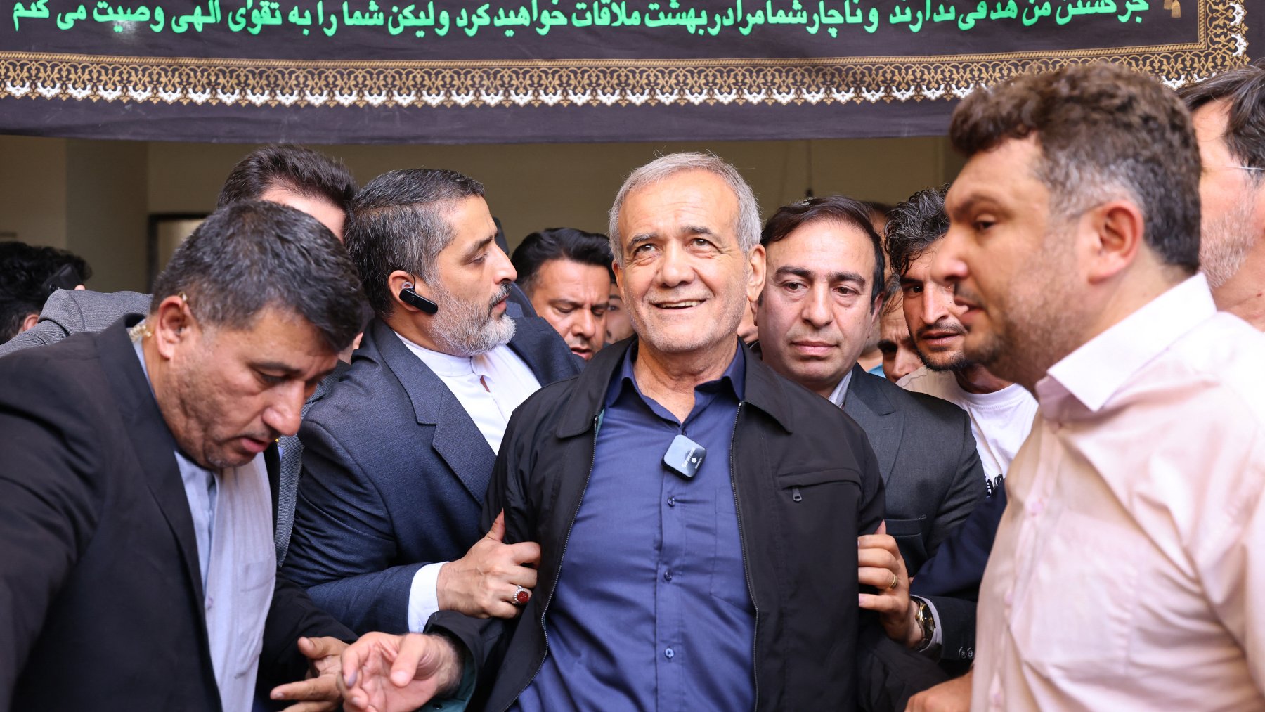 در این تصویر، گروهی از مردان در یک محیط اجتماعی دور یک مرد میانسال جمع شده‌اند. این مرد لبخند بر لب دارد و به نظر می‌رسد در حال برقراری ارتباط با دیگران است. محیط تصویر نشان‌دهنده‌ی یک رویداد مهم یا تجمع است. مردان اطراف او به نظر می‌رسند که به او احترام گذاشته و از او حمایت می‌کنند. متن در بالای تصویر به زبان فارسی نوشته شده است و به نظر می‌رسد حاوی یک پیام یا عنوان خاص باشد.